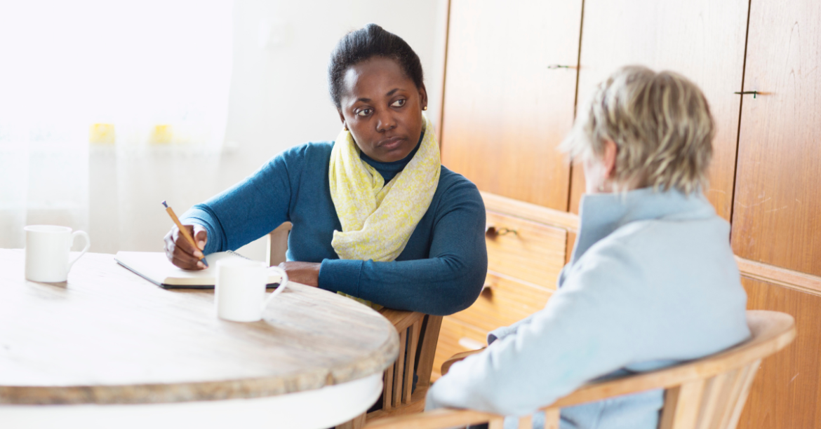 Feature photo of 5 surprising roles of a hospice social worker. Black woman social worker talks with white elderly patient at kitchen table. 